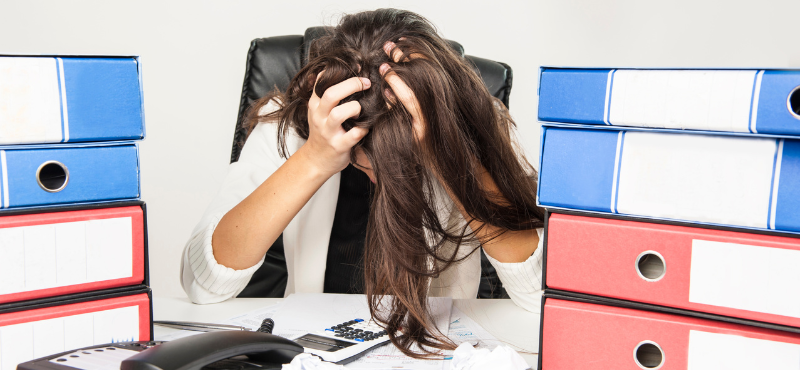 woman running hands through hair, stressed, on a desk with a lot of files and office supplies